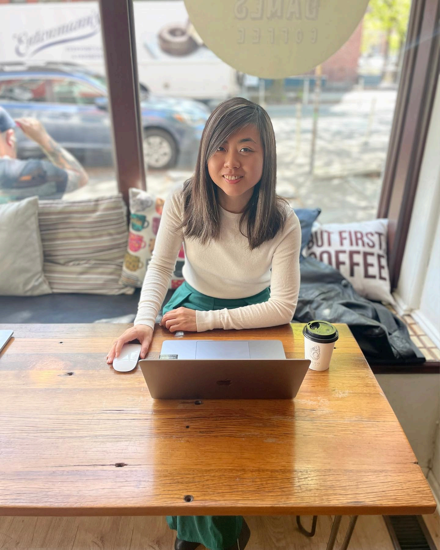 Cherry sitting at a wooden cafe table with a laptop and a coffee cup.