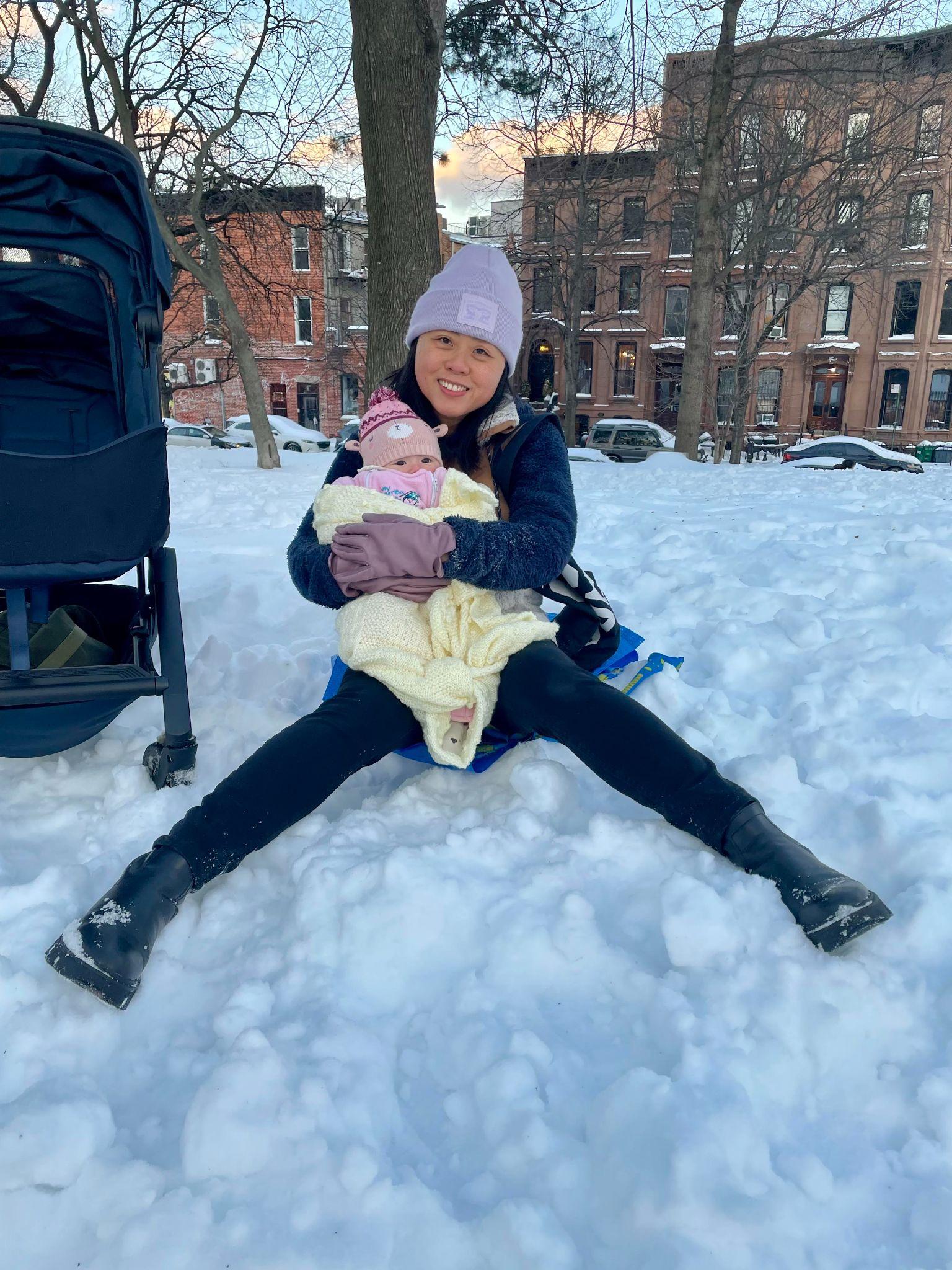 Cherry in a beanie sitting in the snow holding baby Mia wrapped in a yellow blanket.
